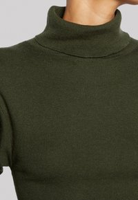 Pull à col roulé vert olive avec une texture côtelée. Manches courtes et coupe ajustée, mettant en valeur le matériau tricoté et la finition lisse.
