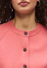 Nő alsó arca fényes ajkakkal, göndör hajjal, arany karika fülbevalóval, rózsaszín gombos pulóvert visel fekete gombokkal.