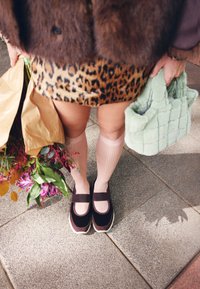 Sapatos de veludo castanho com uma aba, combinados com meias caneladas cor de rosa claro. Também são visíveis uma saia estampada de leopardo, uma mala verde felpuda e um ramo de flores.