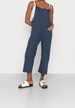 Marineblå dungarees med justerbare stropper, to forsætpunkter og cropped ben-design, parret med hvide slip-on sandaler med sort sål.