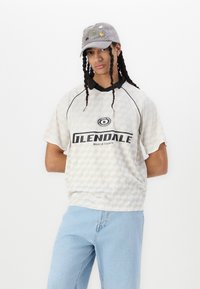 White checkered short-sleeve shirt with black collar, featuring "GLENDALE" text in bold. Grey cap with colorful embellishments. Light blue jeans.