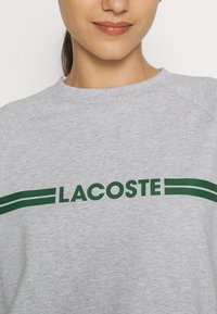 Pilkas džemperis su žaliu "LACOSTE" logotipu ir juostelėmis. Turi apvalią iškirptę ir raglan tipo rankoves. Minkšta tekstūra ir laisvalaikio stilius.