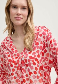 Blusa in tessuto rosa chiaro con scollatura a V e maniche lunghe, caratterizzata da un motivo di cuori rossi e rosa sparsi. Texture liscia.
