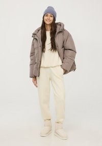 Giubbotto imbottito color taupe, maglione crema e pantaloni crema chiaro; berretto grigio; stivali in morbido tessuto con interno in pelliccia; vestibilità rilassata.