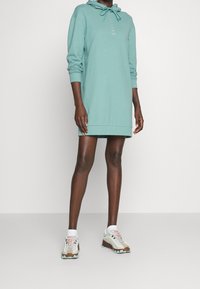 Person som bär en teal långärmad huvtröjklänning med dragsko och vita sneakers med ljusblå och bruna accenter.