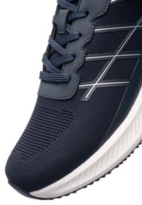 Detailní záběr námořnicky modré sportovní boty s pletenou tkaninou, prošitými překryvy, vzorovanými tkaničkami, reflexními stříbrnými pruhy a bílou polstrovanou podešví.