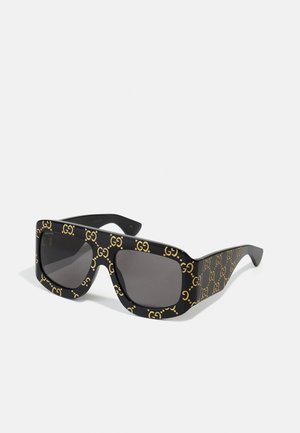 Gafas de sol negras con montura cuadrada de gran tamaño que presenta un patrón repetido del logo dorado "GG". Lentes grises con una textura suave.