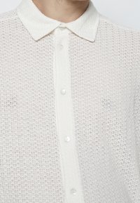 Camisa blanca de botones con un patrón de tejido texturizado y un suave cuello clásico. Presenta botones claros en la parte delantera.