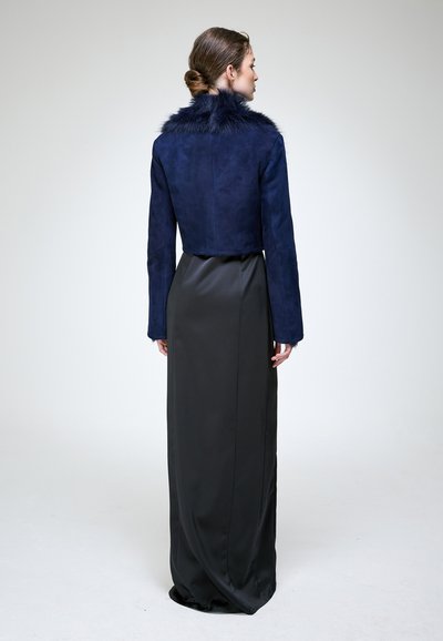 Femme aux cheveux bruns en chignon bas, portant une veste courte bleu marine avec col en fourrure et une longue jupe noire en satin, debout de dos sur un fond uni.