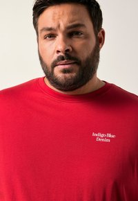 Červené bavlněné tričko s kulatým výstřihem a krátkými rukávy. Na levé části hrudi je vytištěno v bílé barvě 'Indigo Blue Denim'.