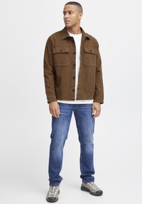 Chaqueta de camisa de algodón marrón con dos bolsillos en el pecho, cierre de botones y corte recto, combinada con jeans azules y zapatillas grises.