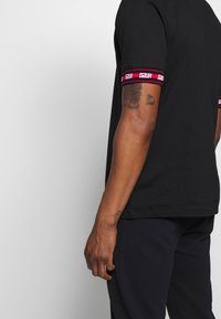Camiseta de manga corta negra con una banda de contraste en rojo y rosa en las mangas, que presenta detalles del logotipo. La tela parece suave y tiene un ajuste relajado.