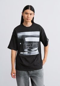 Sort oversized t-shirt med grafisk print og tekst, der viser racingelementer. Blødt stof og afslappet pasform. Bæres med grå jeans.