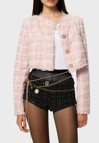Pink tweed cropped jacket with textured grid pattern, featuring gold-tone buttons. Paired with black shorts and layered gold chains.