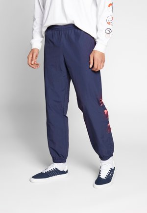 Mężczyzna ubrany w granatowe spodnie dresowe z różowymi geometrycznymi akcentami po bokach, białe skarpetki, granatowo-białe buty sportowe oraz białą koszulę z długim rękawem.