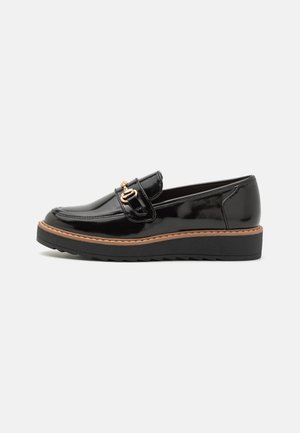 Mocassin en cuir noir brillant avec détail en boucle dorée, bord tan et semelle plateforme noire crantée, présenté en profil latéral sur fond blanc.
