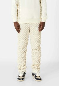 Crèmegekleurde gebreide trainingsbroek met een gestructureerde, gemusterde stof. Aansluitende boorden en een zijlabel. Gecombineerd met zwarte en beige sneakers.
