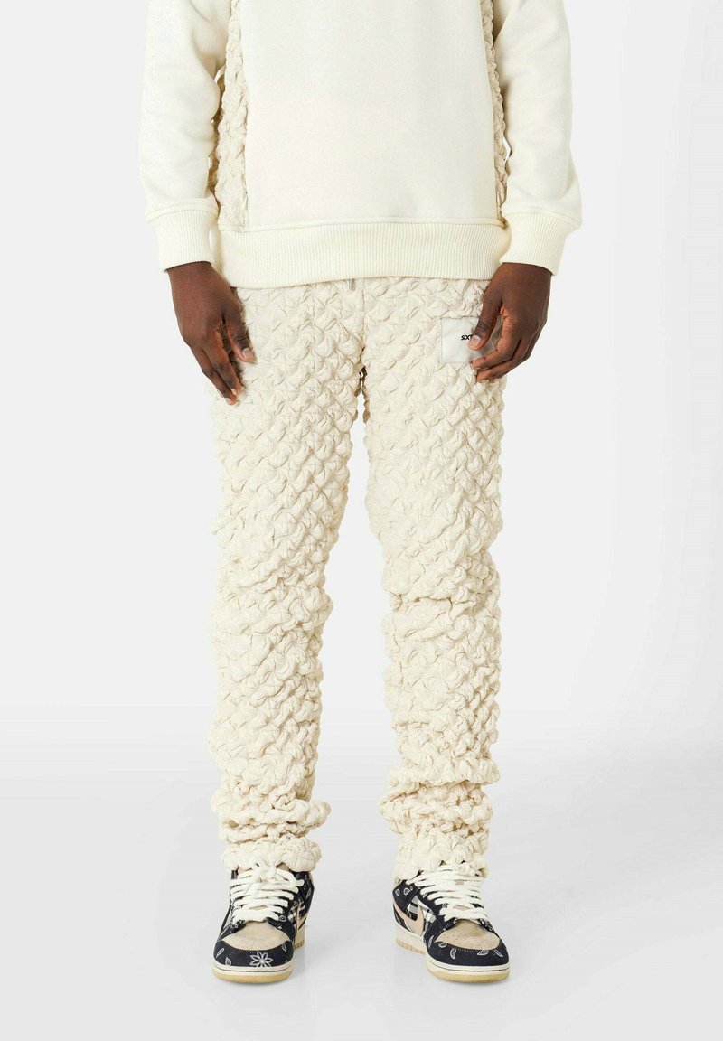 Crèmegekleurde gebreide trainingsbroek met een gestructureerde, gemusterde stof. Aansluitende boorden en een zijlabel. Gecombineerd met zwarte en beige sneakers.