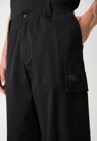 Svarte cargo-bukser har knapp og elastisk liv, sidelommer, og en frontlomme med klaff og logopatch. Knappe glatt stofftekstur.