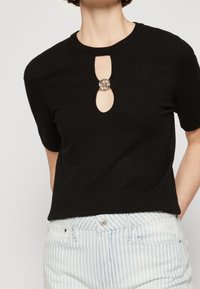 Czarny top z krótkim rękawem, z okrągłym dekoltem i wycięciem w kształcie łezki na środku, ozdobiony okrągłym srebrnym akcentem. Gładka, dzianinowa faktura.