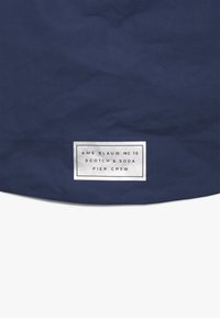 Granatowa tkanina o teksturowanej powierzchni, z białą metką zawierającą napisy "AMS BLAUW N° 16," "SCOTCH & SODA" oraz "PIER CREW."