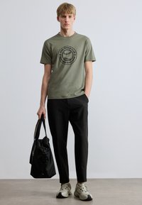 Olívazöld póló fekete kör alakú teniszező klub grafikával, fekete nadrággal és világos színű tornacipővel párosítva, kezében fekete edzőtáskával.