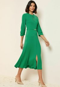 Vestito midi verde con scollatura rotonda, maniche a tre quarti, uno spacco laterale e cintura in vita con hardware argentato. Tessuto dalla superficie liscia.