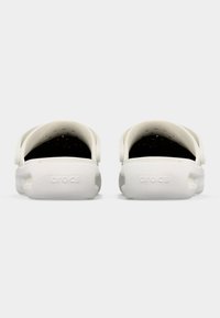 Ζευγάρι λευκών σανδαλιών Crocs, εμφανιζόμενα από την πίσω πλευρά με ιμάντες για τη φτέρνα και οπές αερισμού σε απλό φωτεινό φόντο.