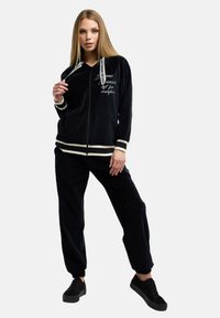 Svart velour-träningsoverall med en zip-up hoodie med vita dragsnören, kontrasterande ränder vid ärmslut och matchande lösa byxor.