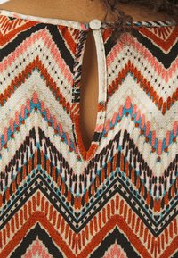 Un tissu texturé présente un motif en zigzag multicolore en orange, rose, noir et bleu, avec une petite fermeture à bouton et un détail en trou de serrure à l'arrière.