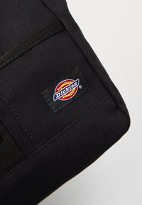Zwarte, textuurstof tas met een hoek waarin een rechthoekig Dickies merklabel is genaaid, in de kleuren rood, geel, blauw en wit.