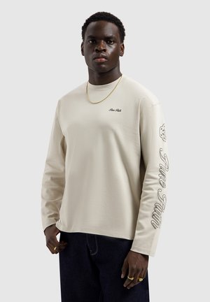 Man draagt een beige shirt met lange mouwen met de tekst 'True Faith' op de mouw en borst, zwarte broek, gouden ketting en ringen.
