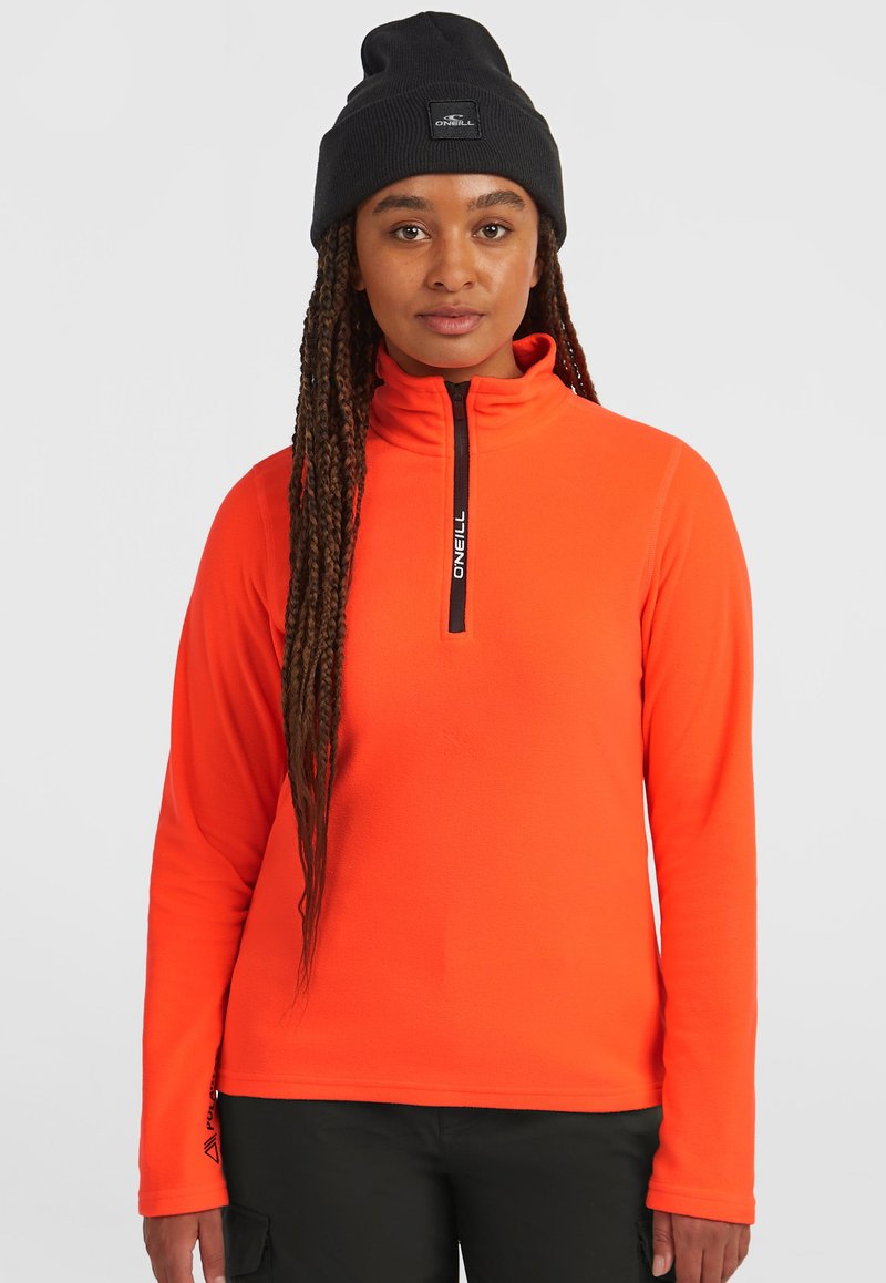 Sudadera de forro polar de color naranja con cremallera y cuello alto; presenta una cremallera negra y un logotipo. Se lleva con un gorro de punto negro, que muestra un acabado texturizado.