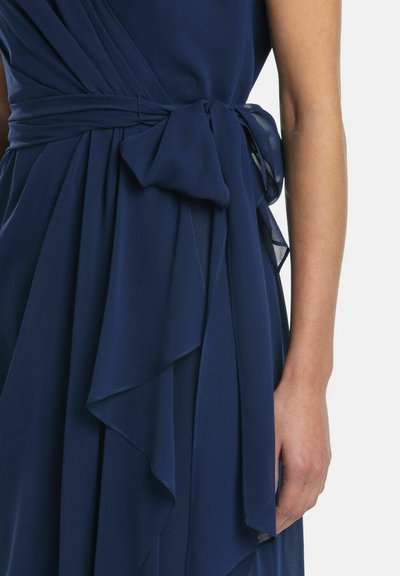 Rochie albastru marin legată în talie cu un arc mare pe partea dreaptă, evidențiind o prim-plan a țesăturii și a unui braț gol.
