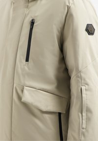 Beige geïsoleerde jas met een matte afwerking, voorzien van een ritsborstzak, een oversized voorzak en een hexagonaal logo patch op de mouw.