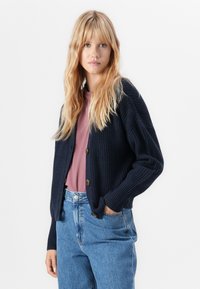 Femme blonde portant un cardigan en maille épaisse bleu marine sur un haut mauve, avec un jeans bleu taille haute, se tenant devant un fond blanc.