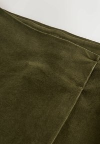 Tuvumā redzams salocīts olīvzaļš kordūra audums, kas parāda mīksto tekstūru un ribotu rakstu uz gaišas virsmas.