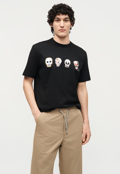 Νέος άντρας φοράει μαύρο μπλουζάκι με τέσσερα πολύχρωμα σχέδια κρανίων στο στήθος και μπεζ παντελόνι με κορδόνι, στέκεται μπροστά σε απλό φόντο.