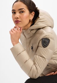 Donna con giacca a piumino beige con cappuccio, appoggiando il mento sulla mano, che indossa piccoli orecchini a cerchio, su sfondo bianco uniforme.