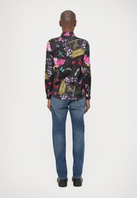 Camicia a maniche lunghe in tessuto nero con motivi colorati di fiori e gabbie per uccelli; abbinata a jeans in denim blu e scarpe nere.