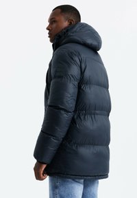 Giacca a vento blu navy con cappuccio, dotata di design trapuntato, texture liscia e una piccola patch con logo sulla manica. Vestibilità casual vista di lato.