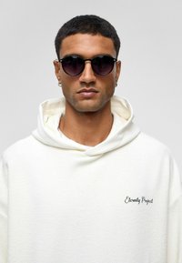 Witte hoodie van structuurstof met zwarte borduursel "Eternity Project". Model draagt ronde zwarte zonnebril.