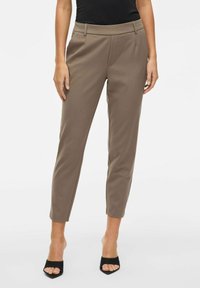 Hnědé kalhoty na míru se slim fit, do kotníků, hladkým povrchem a předním skladem, doplněné černými sandály na vysokém podpatku.