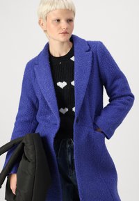 Purple textured, long coat with notched lapels and button closure, layered over a black sweater with white heart patterns, held with a black bag.