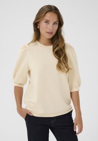 Ljusbeige blus med puffärmar och rund halsringning, tillverkad av mjukt tyg, matchad med mörka jeans. Minimalistisk design, utan mönster.
