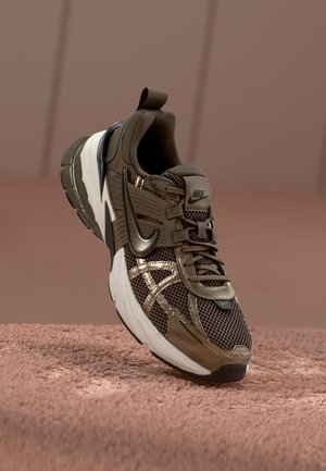 Brązowo-czarny but sportowy Nike z białą podeszwą, cholewką z siateczki i materiału syntetycznego, ustawiony pionowo na teksturowanej brązowej powierzchni.