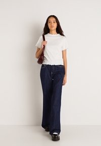 Λευκή μπλούζα με κο scalloped-hem, χαλαρές σκούρες μπλε τζιν παντελόνες και μαύρες πλατφόρμες. Μοντέλο κρατώντας μια καφέ τσάντα χεριού, όρθιο μπροστά από ένα ουδέτερο φόντο.