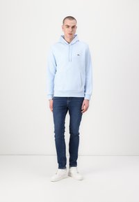 Sudadera de algodón azul claro con un bolsillo frontal tipo canguro, capucha con cordones y un pequeño logo verde. Combinada con jeans oscuros y zapatillas blancas.