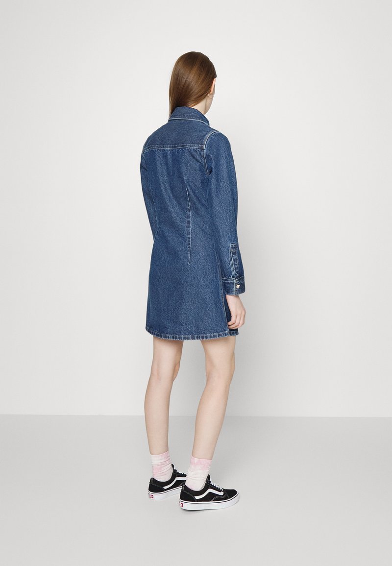Vestido de camisa de mezclilla con collar, mangas largas y silueta ajustada que termina por encima de las rodillas. Combinado con zapatillas negras y calcetines rosas.