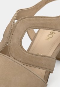 Μπεζ σανδάλι από σουέτ με τετράγωνο τακούνι, με σχέδιο με κοψίματα, ραμμένες λεπτομέρειες και μαλακή υφή στο πάνω μέρος. Χρυσό λογότυπο εσωτερικά.